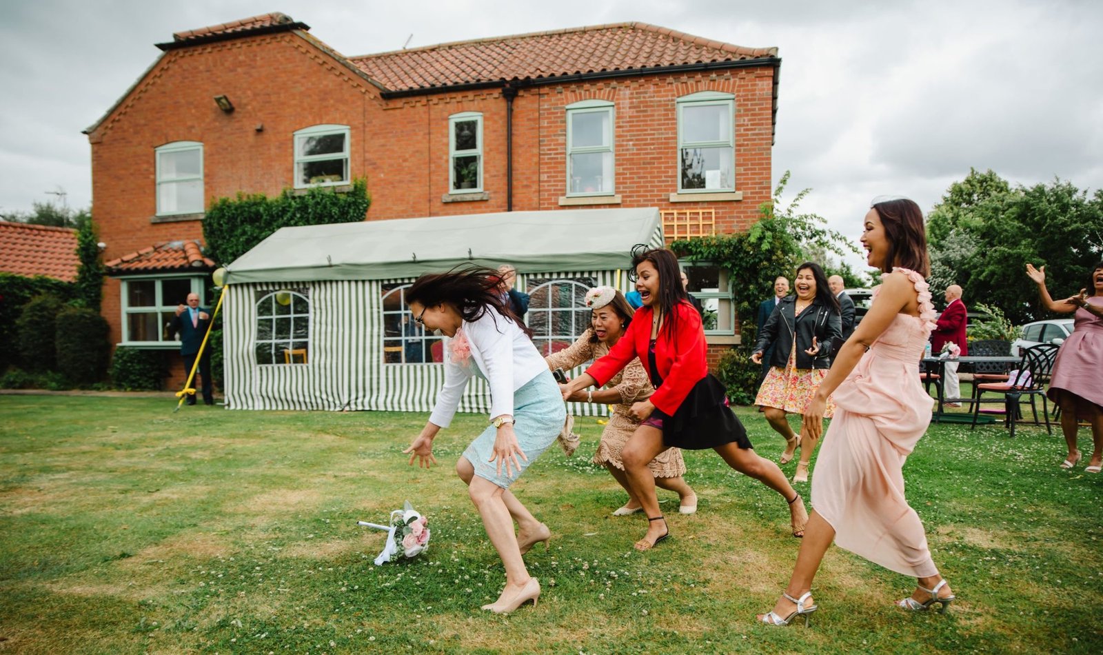 Guests diving for the bouquet on a lawn in front of a red-brick house