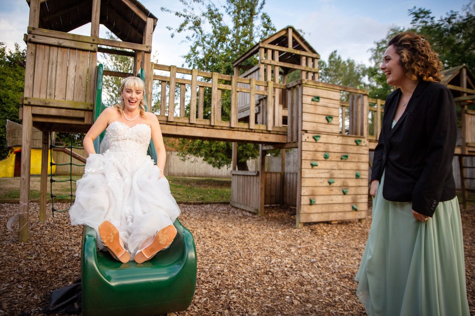 Bride laughing as she comes down a playground slide, a friend watching