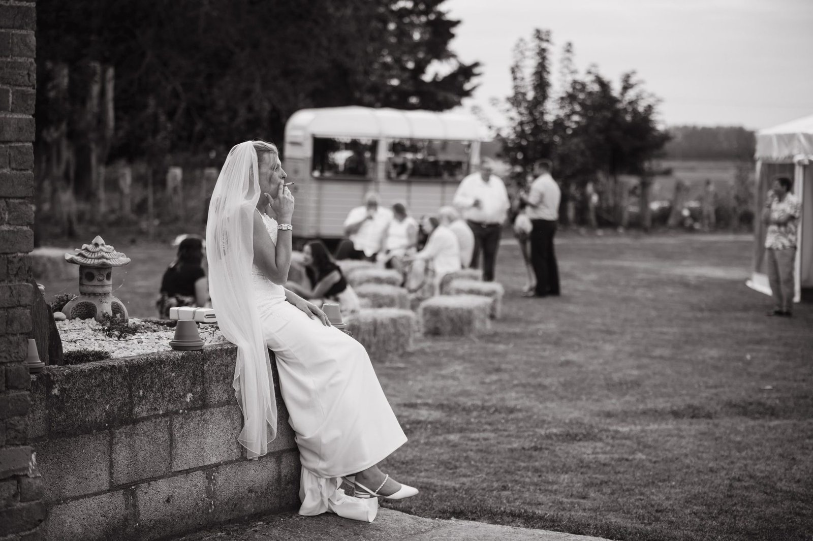 Bride sitting on a garden wall in her veil while guests talk on the lawn, in black and white