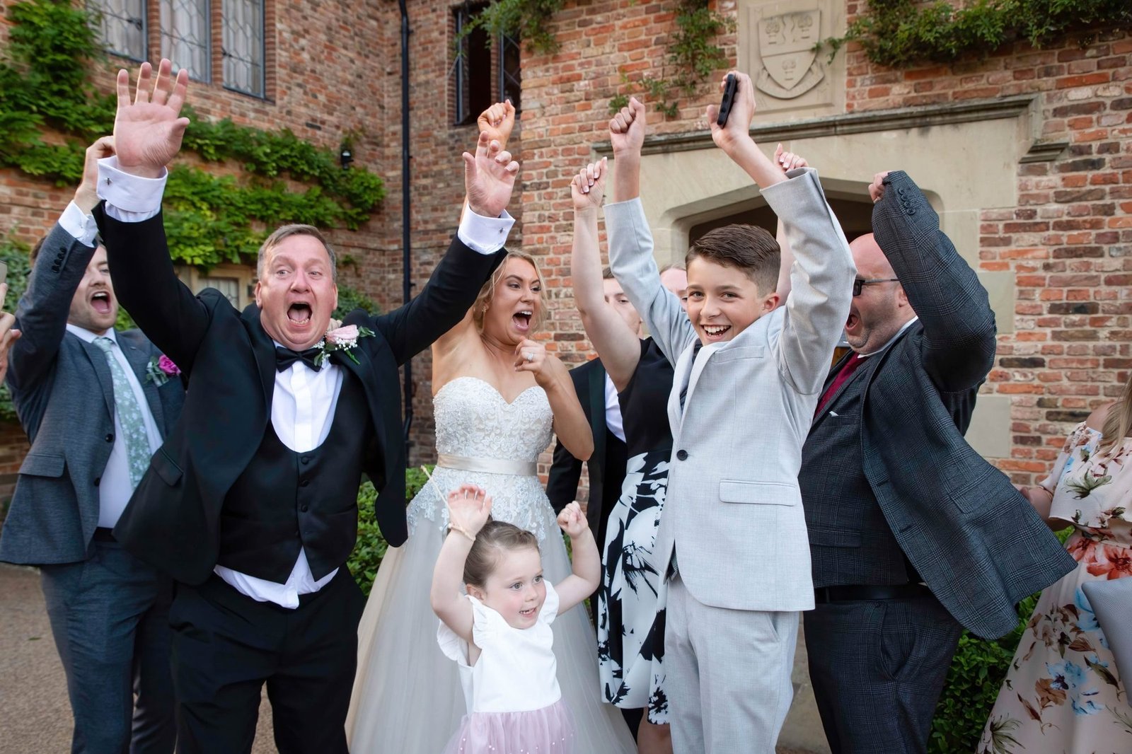 Bride and guests cheering with arms in the air outside a brick country house