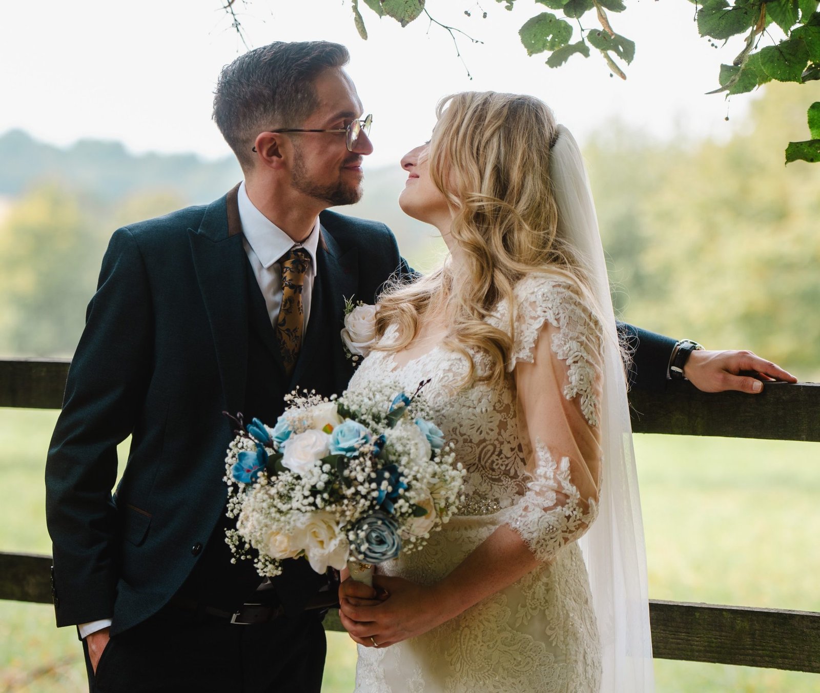 Bride and groom leaning on a fence, looking at each other under the trees