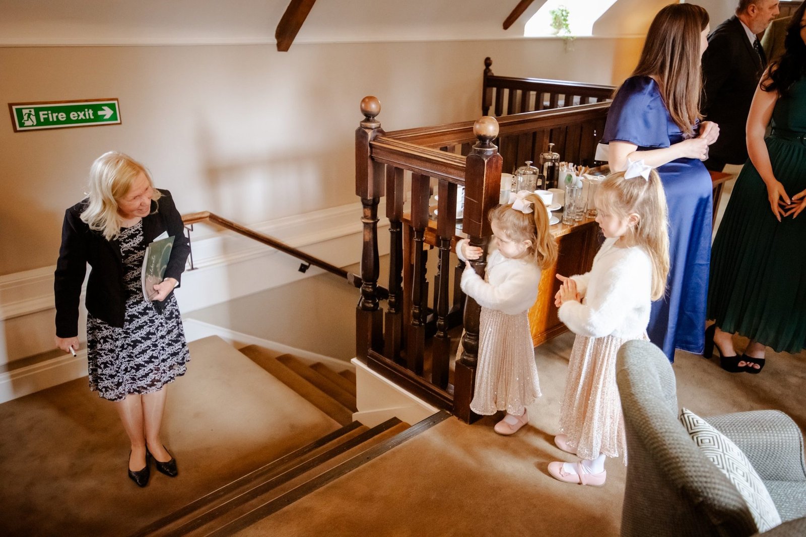 Two flower girls waiting by the banister as the celebrant smiles at them from the stairs